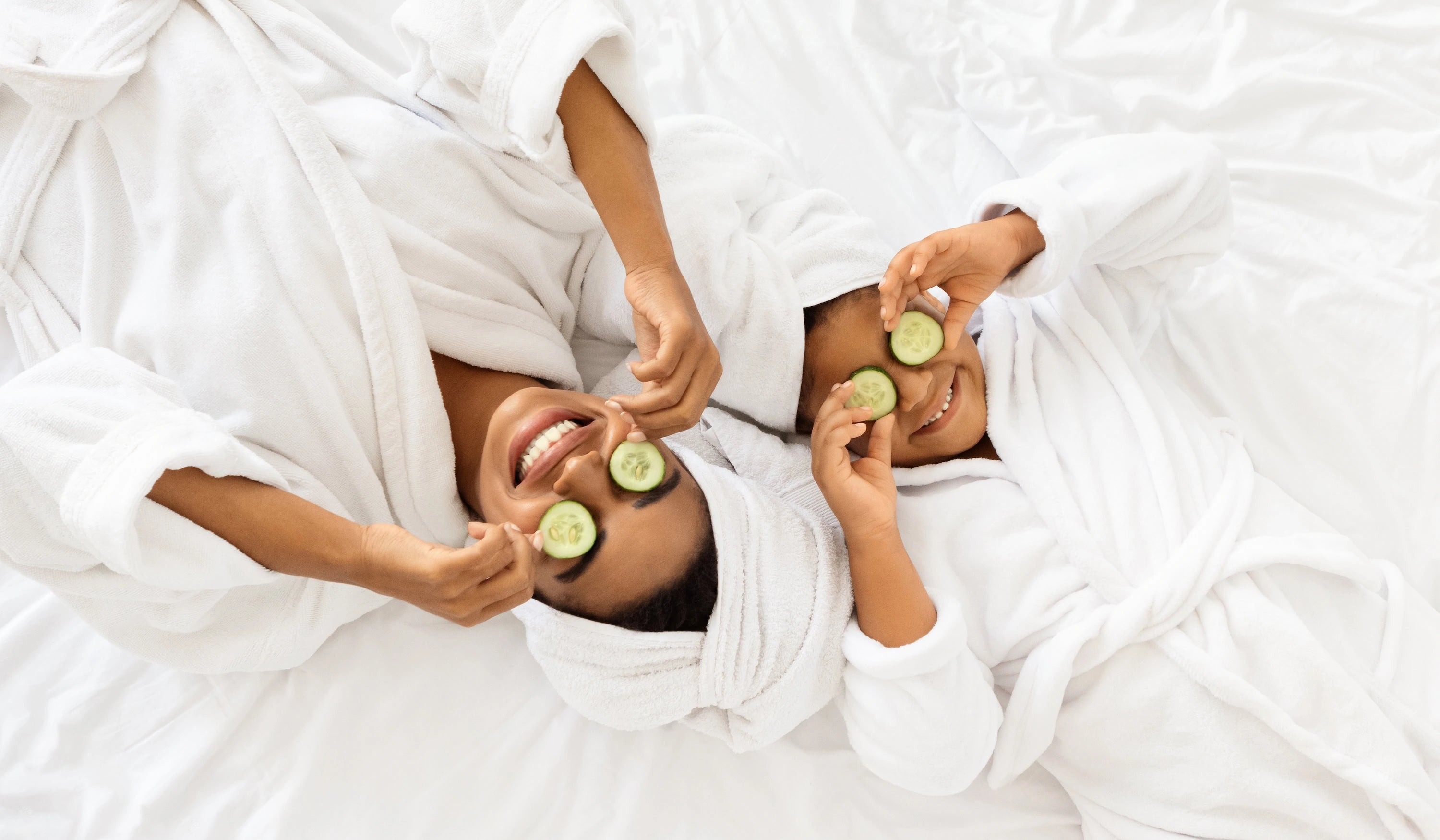Two people lying on a white blanket with cucumbers on their eyes, wrapped in white robes.