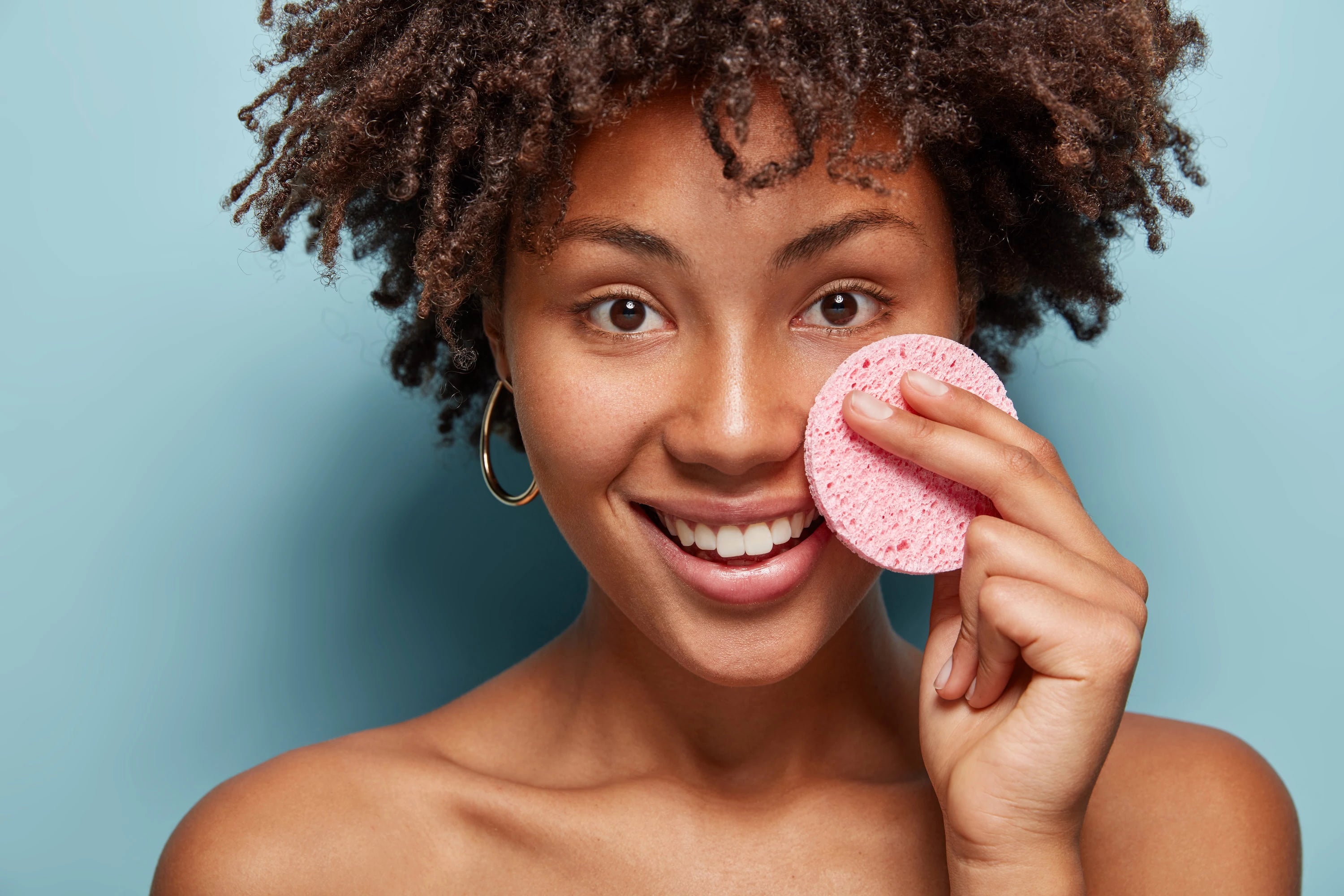 Woman holding a pink sponge against her face with a blue background