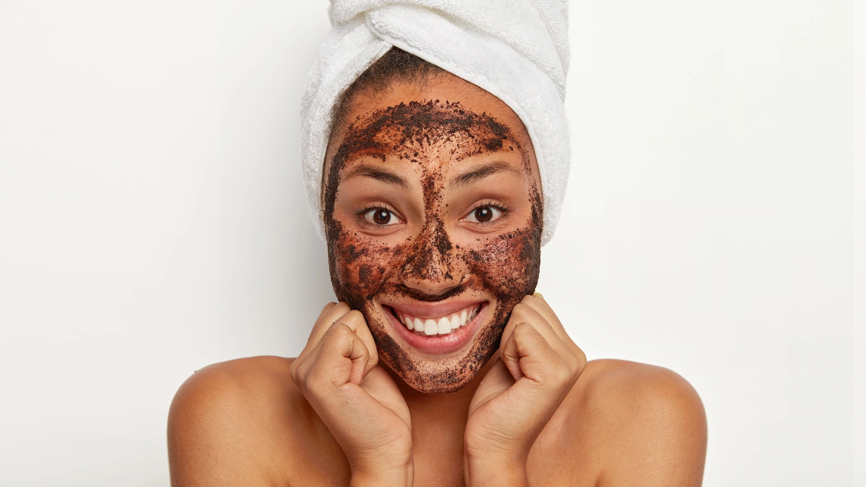 Woman with a coffee face scrub on her face, wearing a towel on her head, against a white background.